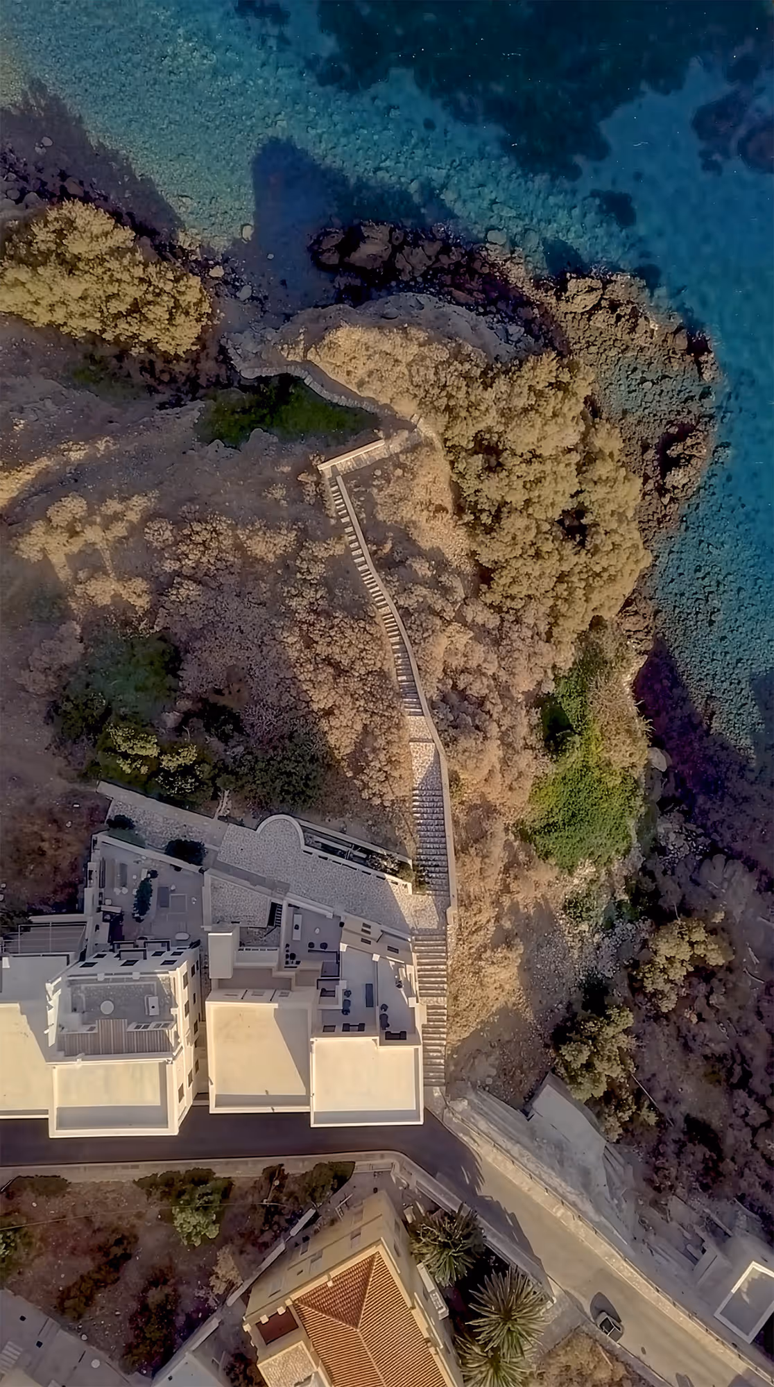Aerial view of coastal buildings, a winding stone staircase, vegetation, and clear blue sea with rocky shore.