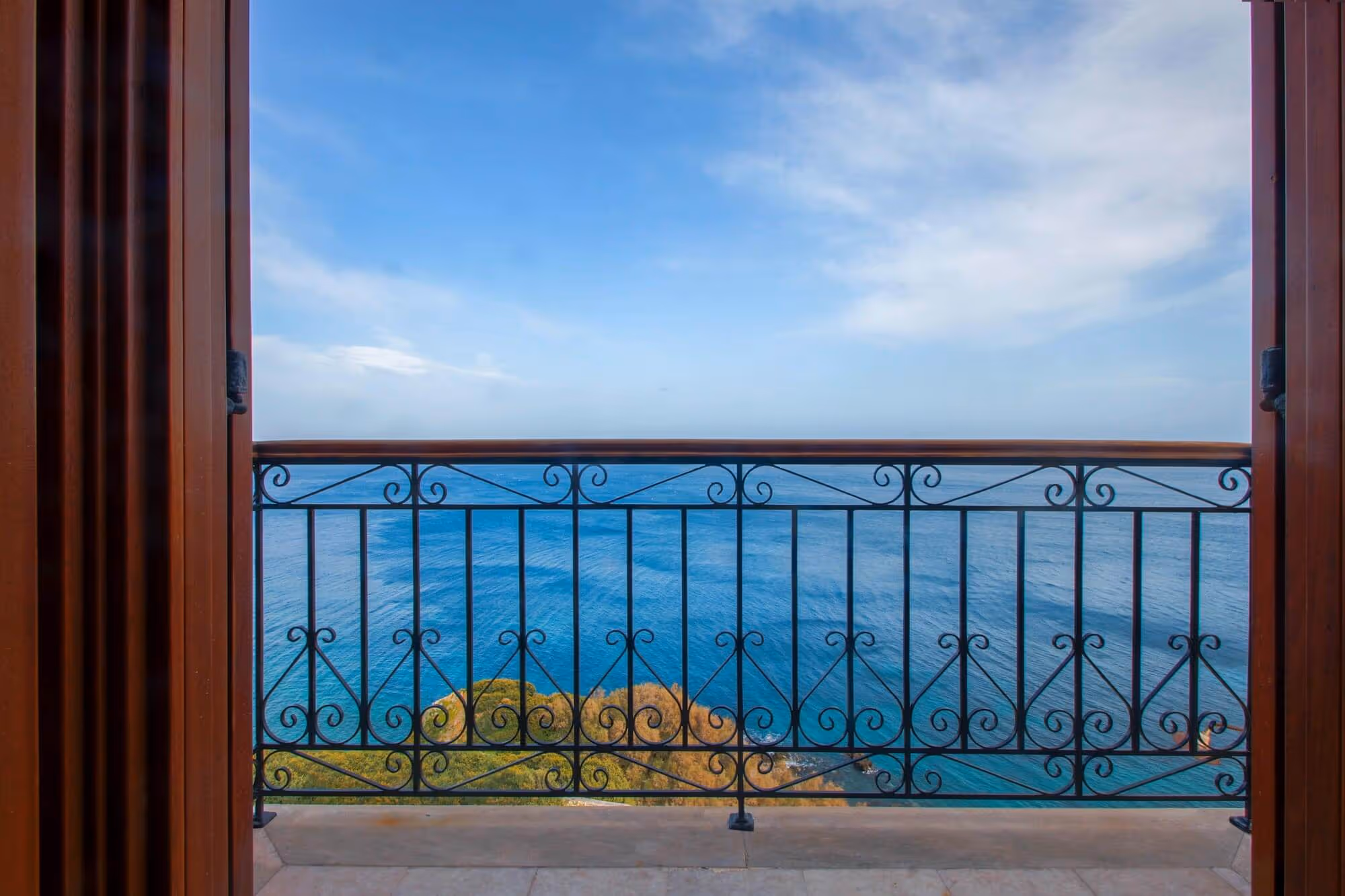View of a blue ocean and partly cloudy sky seen through an open balcony door with ornate black metal railing and wooden frames.