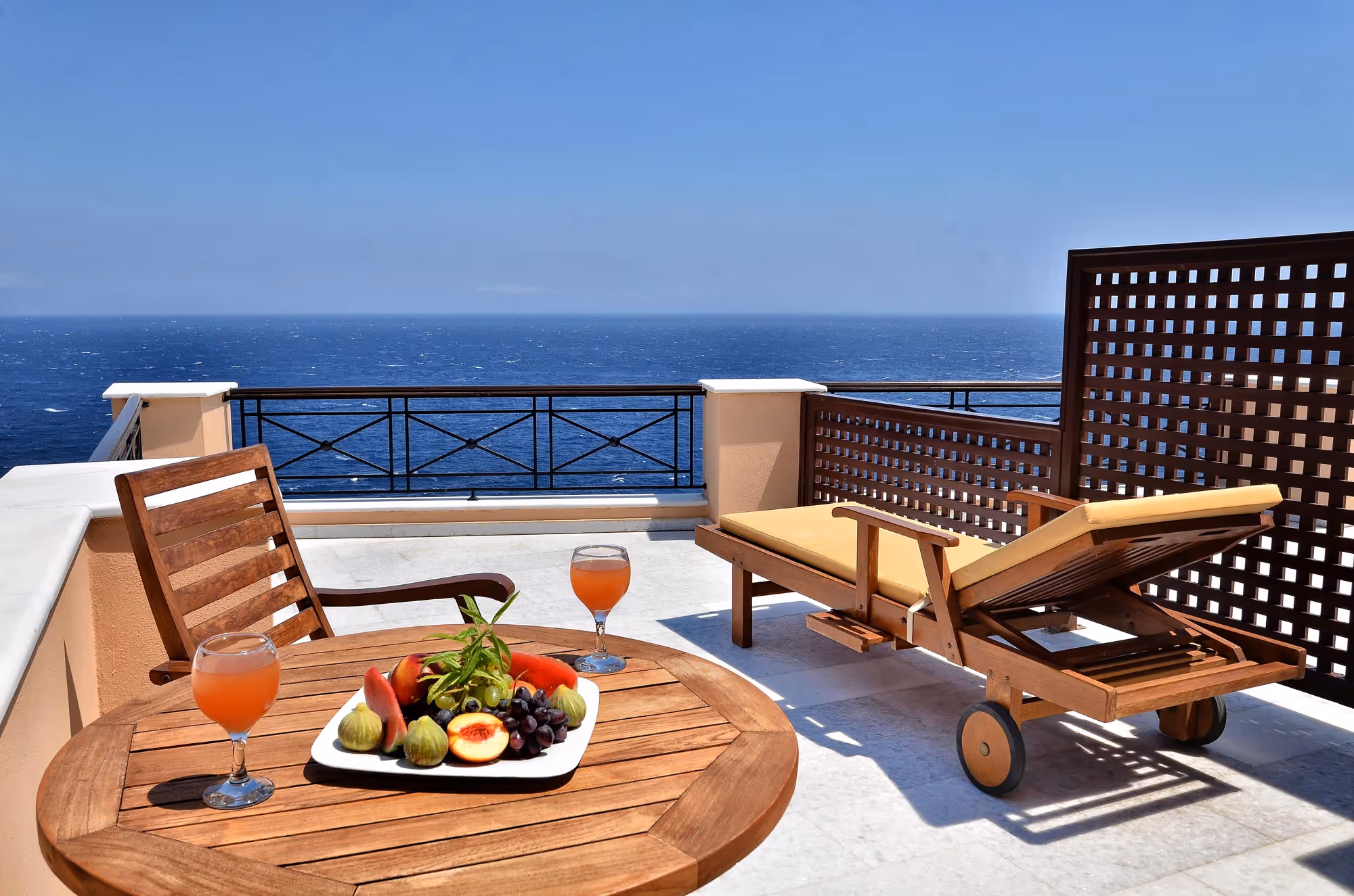 Sunlit terrace overlooking the ocean with a wooden table holding a fruit platter and two glasses of juice, and a wooden lounge chair with yellow cushion.