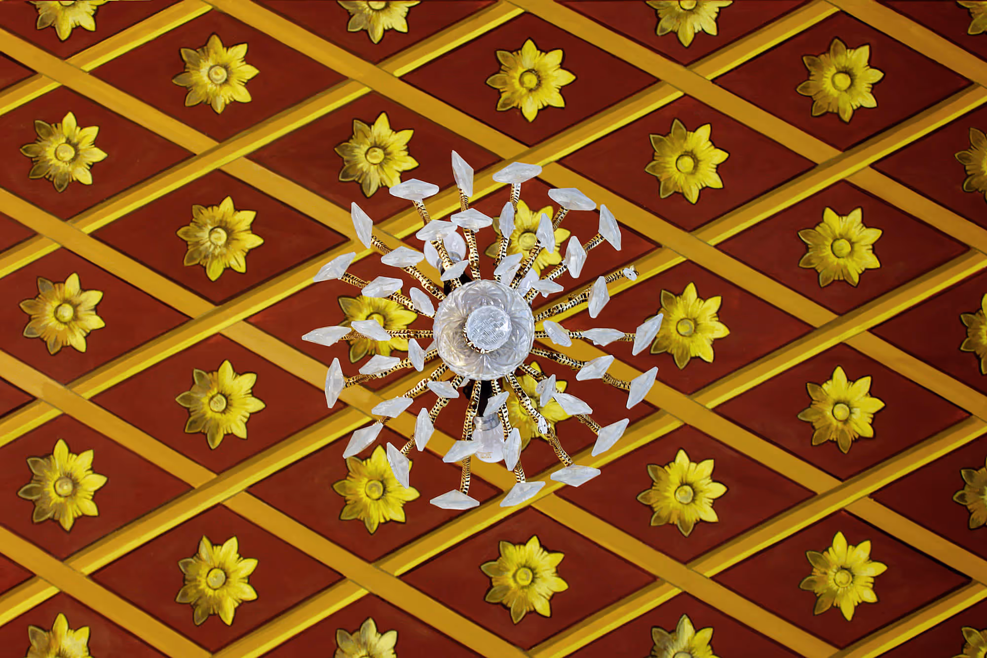 Ornate ceiling with red and gold diamond pattern and yellow floral decorations, centered by a decorative chandelier with multiple glass elements.