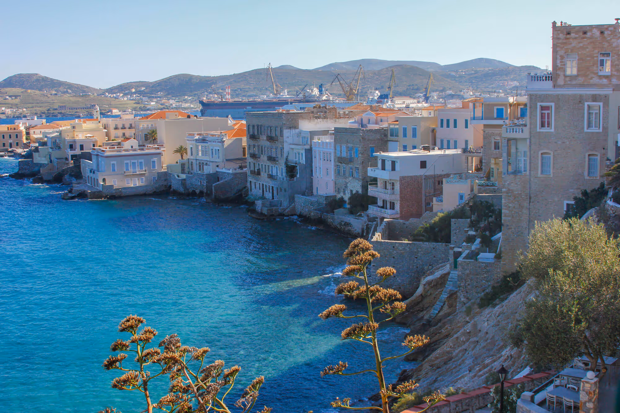 Coastal town with stone buildings and clear blue sea under a clear sky, with hills and a shipyard in the background.