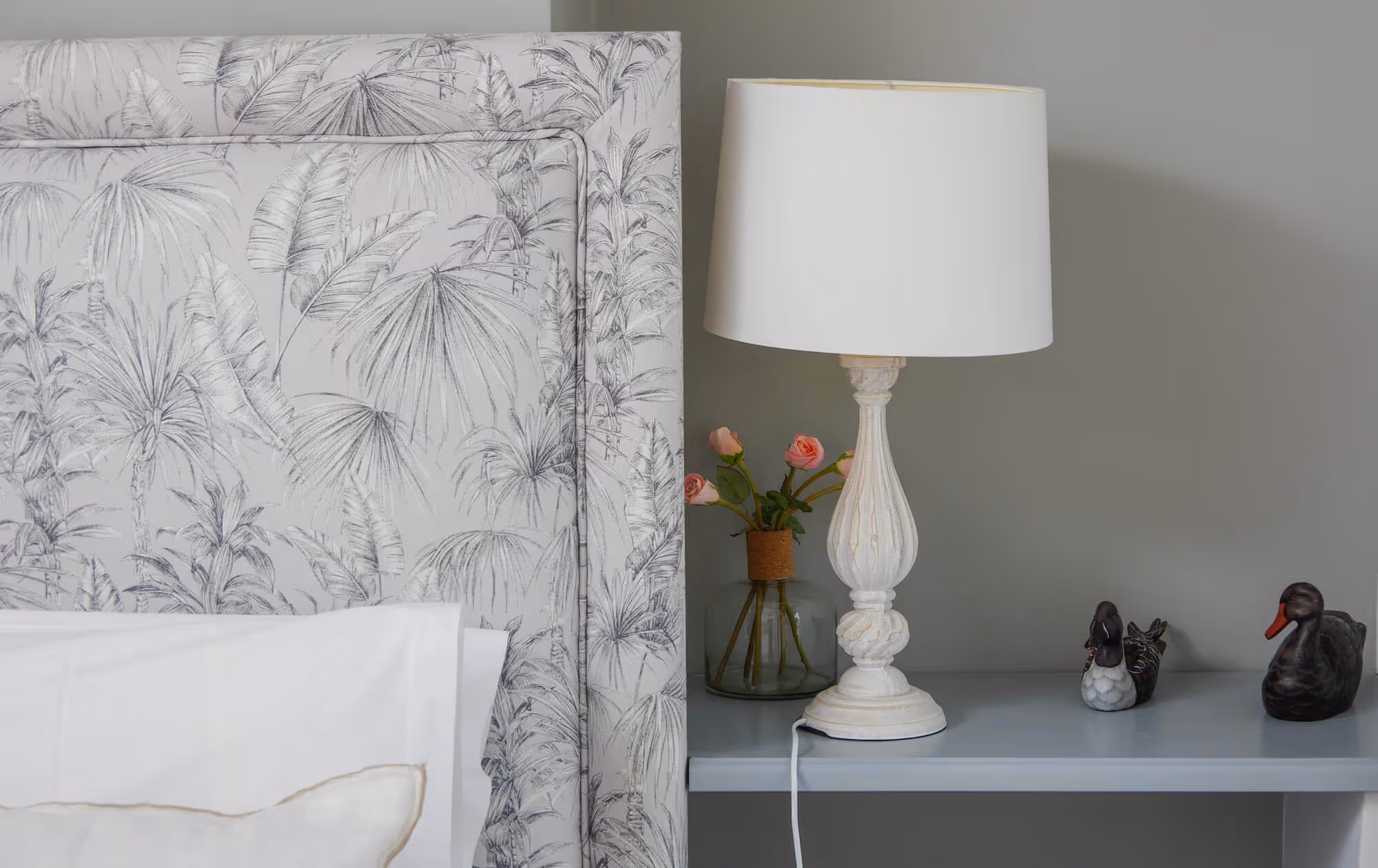 Nightstand with a white decorative lamp, a vase with pink roses, and two black duck figurines next to a bed with a patterned headboard.