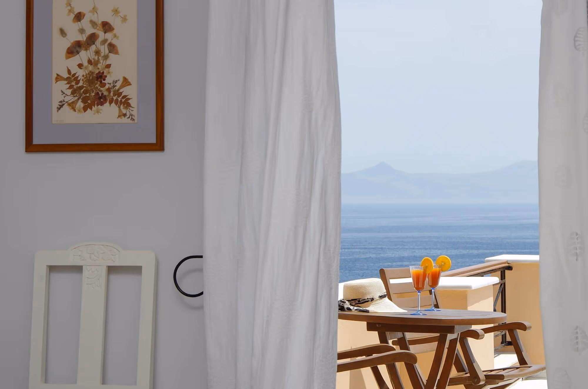 Wooden table and chairs on a balcony with two cocktails garnished with orange slices, overlooking a calm blue sea and distant mountains, partially framed by white curtains.