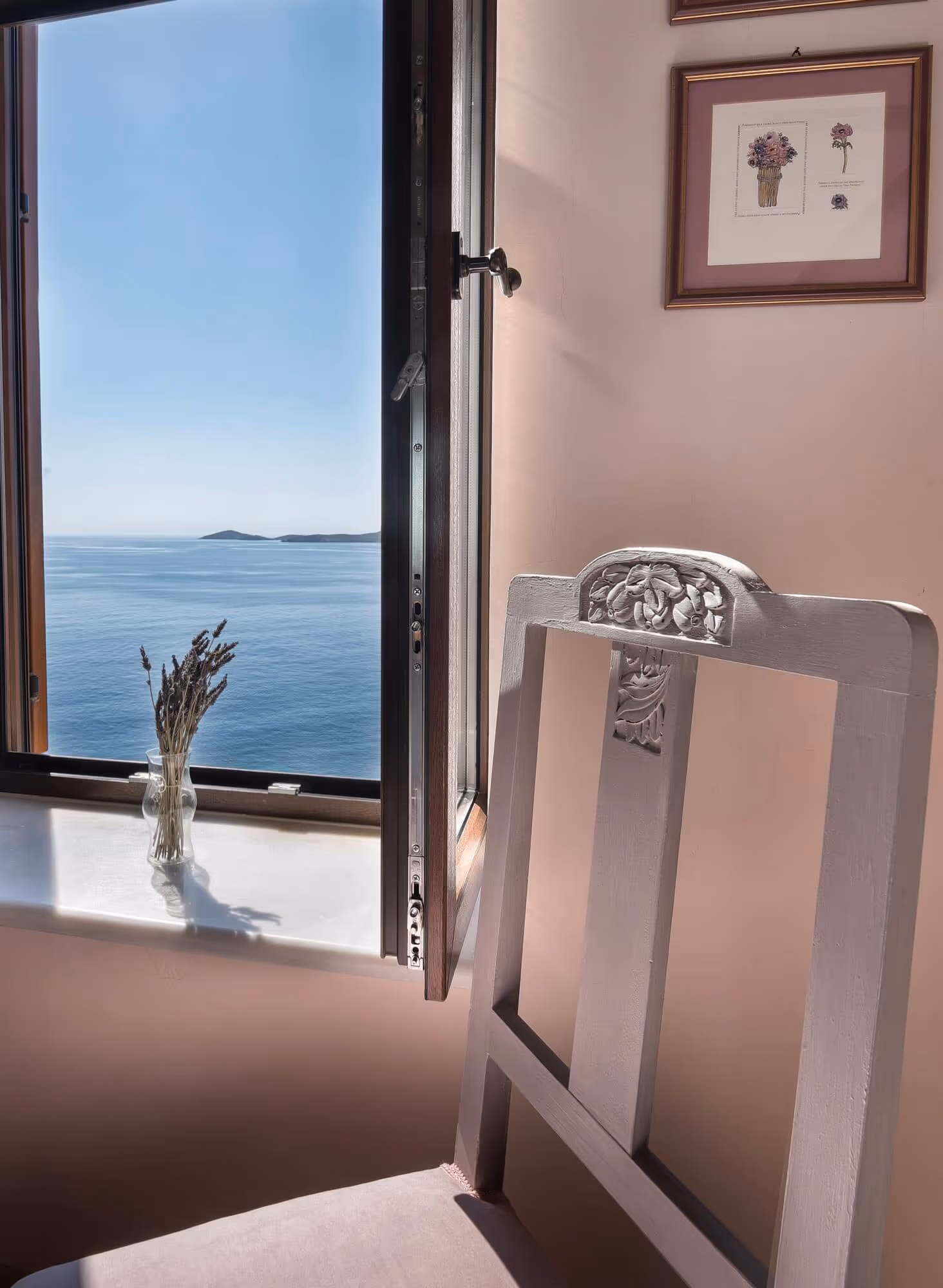 Open window overlooking calm blue sea with a small vase of dried flowers on the windowsill and a carved white chair in soft sunlight.